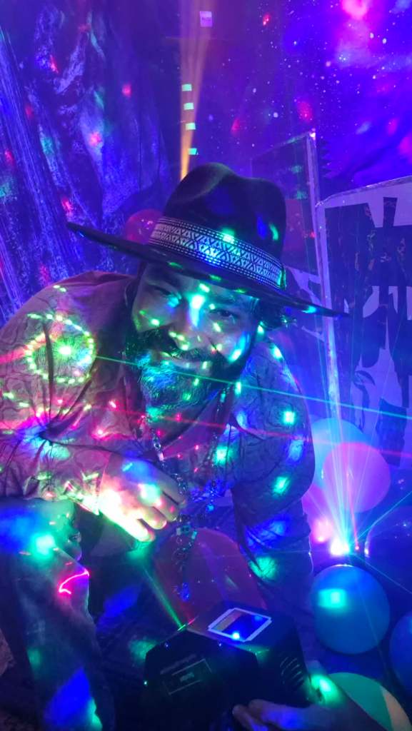 A person with a beard is smiling while surrounded by colorful lights and laser effects, with a party atmosphere in the background.